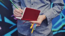 a man working on a logo design makes notes in a small red booklet, he is holding colouring pencils, wearing a denim shirt and standing in front of a stylised colourful background - header image from the graphic design blog by nstudio