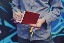 a man working on a logo design makes notes in a small red booklet, he is holding colouring pencils, wearing a denim shirt and standing in front of a stylised colourful background - header image from the graphic design blog by nstudio