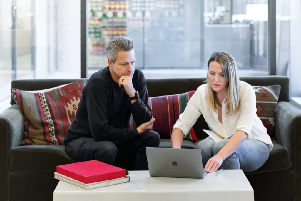 a man and a woman discuss a project on a sofa with a macbook computer