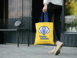 a woman walks down the street holding a yellow happy hounds branded tote bag