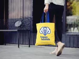 a woman walks down the street holding a yellow happy hounds branded tote bag