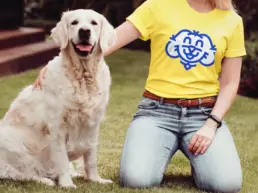 a woman kneels down next to a golden retriever, wearing a yellow happy hounds t shirt. The logo is large on the front side.