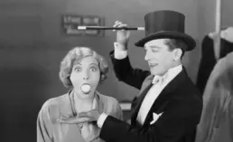 a vintage black and white photograph of a magician and his female assistant, who is surprised to have an have an egg in her mouth
