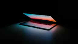 An Apple MacBook laptop is switched on and partly open, with the bright colours lighting the dark scene