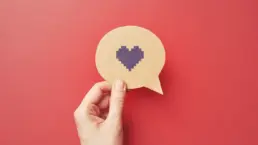 a hand holds a cut out of a speech bubble with a pixel heart icon drawn on it