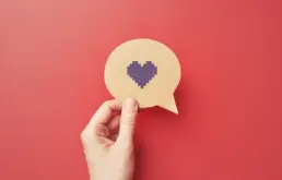 a hand holds a cut out of a speech bubble with a pixel heart icon drawn on it