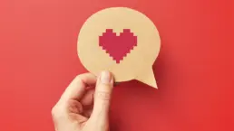 a hand holds a cut out of a speech bubble with a pixel heart drawn on it