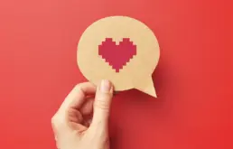 a hand holds a cut out of a speech bubble with a pixel heart drawn on it