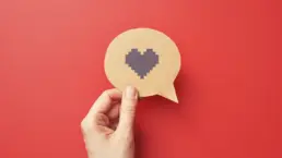 a hand holds a cut out of a speech bubble with a pixel heart icon drawn on it