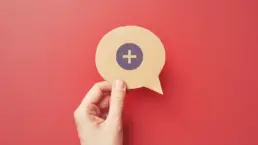 a hand holds a cut out of a speech bubble with a plus icon drawn on it